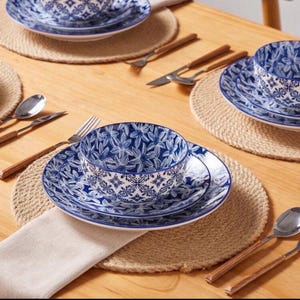 Puede incluir: Un juego de vajilla de cerámica con un patrón floral azul y blanco, que incluye un tazón apilado sobre un plato. Las piezas están dispuestas sobre manteles individuales redondos tejidos con cubiertos de acero inoxidable y mangos de madera.