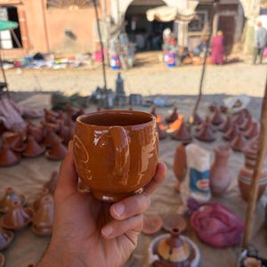 Puede incluir: Una taza de cerámica marrón con asa se sostiene frente a una exhibición de mercado. La taza está rodeada de otros artículos de cerámica, incluyendo tajines y pequeñas ollas. El fondo muestra un entorno de mercado con tiendas y edificios.