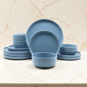 Puede incluir: Un juego de vajilla de cerámica azul claro que incluye varios platos y cuencos redondos apilados en un grupo. Las piezas presentan un acabado liso y mate con bordes simples y limpios, dispuestos en una composición equilibrada.