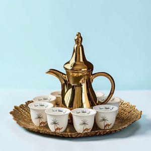 以下が含まれることがあります： 高い蓋と湾曲したハンドルを備えた光沢のある金色のコーヒーポットと、ヤシの木とラクダのイラストが描かれた6つの白いセラミックカップが、質感のある金色の円形サービングトレイの上に配置されています。