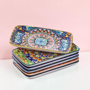 Puede incluir: Una pila de cinco bandejas rectangulares de cerámica con un patrón vibrante y multicolor que presenta intrincados motivos florales y geométricos en tonos azul, amarillo, naranja y verde, con un diseño central tipo mandala en cada una.