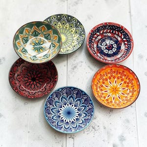 Könnte beinhalten: Ein Set aus fünf runden Keramikschalen mit komplexen, farbenfrohen Mustern im Mandala-Stil. Die Schalen zeigen verschiedene Farbpaletten, darunter Grün und Gelb, Dunkelrot und Schwarz, Dunkelblau und Weiß, leuchtendes Orange und Gelb sowie Blau.