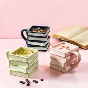 Puede incluir: Tres tazas de cerámica cuadradas con forma de pilas de libros. Una taza es azul oscuro con hielo, una es verde claro con una rodaja de limón y menta, y una es rosa claro con trozos de fresa. Cada taza cuenta con un asa y diseño de lomo.