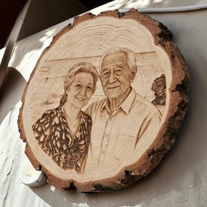 Puede incluir: Un rodaja de madera circular con un retrato pirograbado de una pareja mayor. La veta natural de la madera es visible en toda la pieza, con la corteza conservada en los bordes exteriores. El arte detallado muestra a una persona con cabello largo y una blusa estampada junto a una persona con cabello corto y camisa de cuello, sobre un fondo de playa.