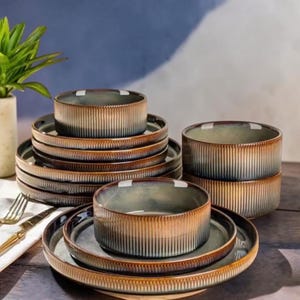 Op de afbeelding: Een set keramisch servies met een geribbelde textuur en een kleurverloop dat overgaat van een diep blauwgrijs midden naar een warme, aardse bruine rand. De collectie bevat gestapelde ronde borden en diepe kommen met een consistent ontwerp.