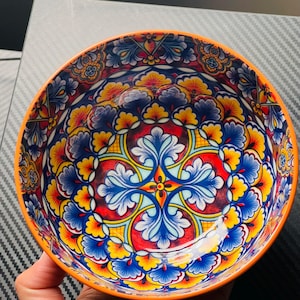 Puede incluir: Un cuenco de cerámica decorativo con un vibrante estampado floral. El cuenco presenta un borde naranja brillante y un diseño detallado en rojo, azul, amarillo y blanco. El cuenco se sostiene en una mano.