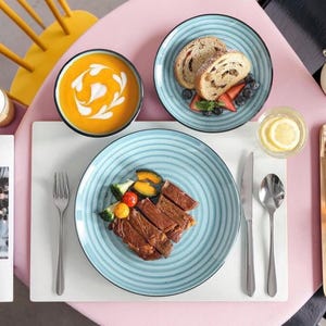 May include: A table setting featuring a light blue ribbed ceramic plate with sliced steak and vegetables, a matching plate with bread and berries, and a bowl of orange soup with cream swirls. Stainless steel cutlery sits on a white rectangular placemat.