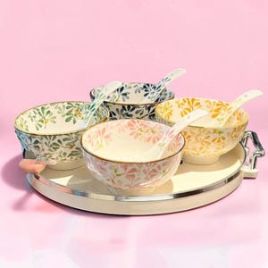 Puede incluir: Un juego de cuatro cuencos de cerámica con cucharas a juego sobre una bandeja redonda blanca. Cada cuenco presenta un patrón floral distinto en verde, azul, rosa o amarillo con bordes dorados. Las cucharas son blancas con detalles florales.