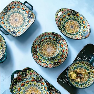 Puede incluir: Un juego de vajilla de cerámica con un patrón repetitivo inspirado en mandalas en tonos verde, naranja, amarillo y azul. El conjunto incluye platos cuadrados y ovalados con asas, cuencos, platos y una bandeja oscura con cubiertos dorados.