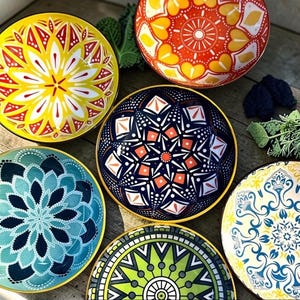 Puede incluir: Un conjunto de seis cuencos redondos de cerámica con intrincados y coloridos patrones estilo mandala en amarillo, naranja, azul oscuro, verde azulado, verde y crema. Cada cuenco muestra un diseño geométrico único con motivos radiales.