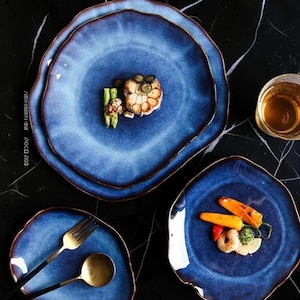 Puede incluir: Un juego de platos de cerámica con formas orgánicas irregulares, esmalte azul degradado profundo y bordes de color marrón oscuro. Los platos tienen un acabado reactivo y se presentan con cubiertos dorados y porciones de verduras y mariscos.
