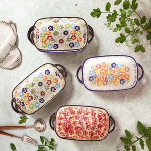 Könnte beinhalten: Vier rechteckige Butterdosen aus Keramik mit abgerundeten Ecken und seitlichen Griffen. Jede Dose zeigt ein einzigartiges, buntes Blumenmuster in Rot-, Blau-, Gelb- und Grüntönen auf cremefarbenem Grund mit dunklen Rändern.