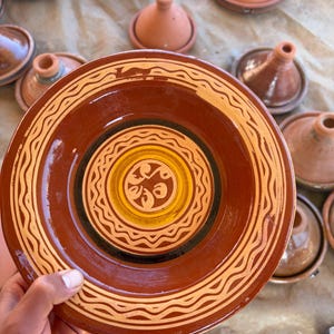 Puede incluir: Un plato de tagine de cerámica marrón con un borde decorativo y un diseño central en amarillo, negro y crema. Otros platos de tagine y tapas son visibles en el fondo. El plato está sostenido por una mano.