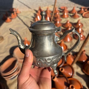 Puede incluir: Una tetera plateada vintage con detalles ornamentados, un pico curvo y un asa pequeña. La tetera se sostiene en una mano, con un fondo borroso de tajines y platos de terracota. La tetera tiene tres patas pequeñas.