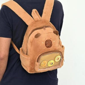 Puede incluir: Una mochila de felpa marrón con un diseño de cara de oso. La mochila tiene un pequeño bolsillo frontal con una ventana transparente que muestra tres pines redondos. Las correas y el cuerpo de la mochila son de un material suave y peludo.
