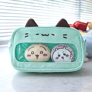 Puede incluir: Un bolso bandolera en forma de gato de color verde azulado claro con orejas marrones y rasgos faciales bordados. El bolso tiene una ventana transparente que muestra dos personajes de peluche. Uno es una bola de masa hervida beige con una cara sonriente, y el otro es un botón rosa y blanco con el texto "RYOUKAI".