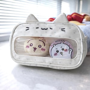 以下が含まれることがあります： 透明な窓が付いた白い猫型のペンケース。ぬいぐるみと丸いピンが入っています。ケースは水玉模様で、猫の耳のアクセントが付いています。ピンには「RYOUKAI」の文字が入った漫画のクマのデザインが施されています。