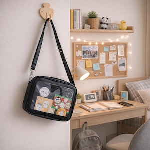 May include: Black crossbody bag with a clear front panel, displaying colorful stickers and pins. The bag hangs from a wooden wall hook. The interior of the bag is visible, showcasing various decorative items.