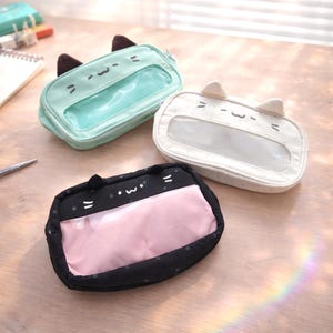 Op de afbeelding: Drie etuis in de vorm van een kat, in mintgroen, wit en zwart. Elke etui heeft een doorzichtig venster en kattenoren. De mintgroene etui heeft bruine oren, de witte etui heeft witte oren en de zwarte etui heeft roze binnenkant.
