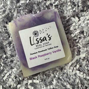 Puede incluir: Una pastilla de jabón artesanal con un diseño de remolino morado y blanco. La etiqueta dice "Lissa's REAL SOAP" y "Black Raspberry Vanilla, 127.5 g". El jabón está sobre un lecho de papel blanco triturado.
