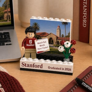 May include: A Lego-style figurine wearing a maroon Stanford sweatshirt stands in front of a miniature scene of a university building. The base reads "Stanford Graduated in 2026." A sign says "Watch Out World, Here I Come!"