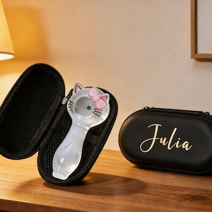 Op de afbeelding: Een heldere, katvormige glazen pijp met een roze strik, in een zwart etui met ritssluiting. Een zwart ovaal etui met de naam "Julia" in gouden script is ook aanwezig. De items staan op een houten oppervlak.