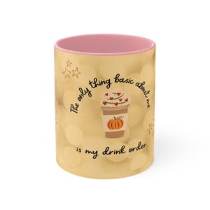 May include: A ceramic coffee mug with a light yellow exterior and a pink interior. The mug features a graphic of a pumpkin spice latte with hearts and a pumpkin. Text on the mug reads, "The only thing basic about me is my drink order."
