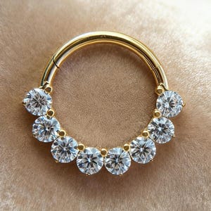 May include: A gold-colored septum ring featuring a series of clear, round-cut gemstones. The gemstones are evenly spaced along the curved surface of the ring, creating a sparkling, circular design. The ring is set against a soft, neutral-toned background.