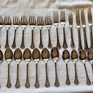 May include: A selection of vintage silverware, including forks, spoons, and knives, arranged on a white cloth. The silverware has a shiny, silver finish and a decorative handle design. The image showcases a complete set of dining utensils.
