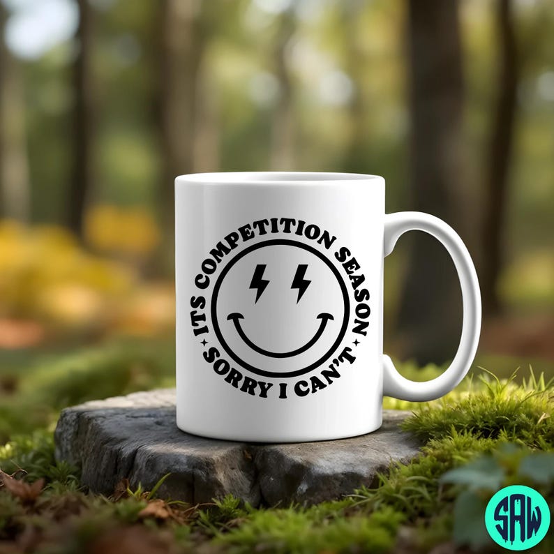 May include: White ceramic mug with a black graphic of a smiley face with lightning bolts and the text "ITS COMPETITION SEASON SORRY I CAN'T". The mug has a curved handle and is sitting on a stone.