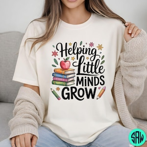 May include: A cream-coloured t-shirt featuring the phrase "Helping Little Minds Grow" in black script. The design includes books, an apple, pencils, and floral elements. The theme is education.