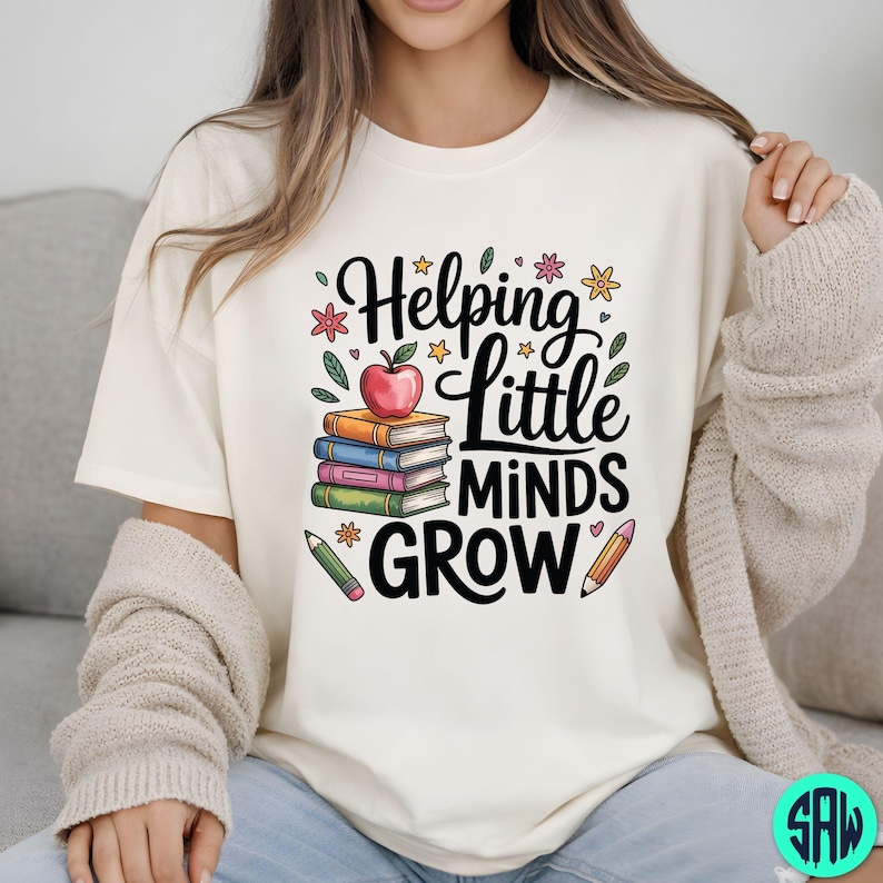 May include: A cream-coloured t-shirt featuring the phrase "Helping Little Minds Grow" in black script. The design includes books, an apple, pencils, and floral elements. The theme is education.