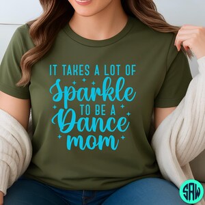 May include: Olive green t-shirt with the text "IT TAKES A LOT OF Sparkle TO BE A Dance mom" in turquoise script. The shirt has a crew neck and short sleeves. The text is surrounded by small star graphics.