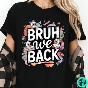 May include: Black t-shirt with the text "BRUH we BACK" in white, featuring cartoon ducks, books, and school supplies. The design includes colourful accents like flowers, hearts, and stars, creating a playful back-to-school theme.