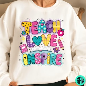 May include: White sweatshirt featuring the words "TEACH LOVE INSPIRE" in a vibrant, cartoonish style. The design incorporates hearts, flowers, an apple, a pencil, and a book, ideal for teachers or those in education.