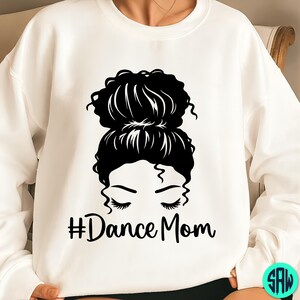 May include: White sweatshirt with a black graphic of a woman's face with closed eyes and a high bun hairstyle. The text "#Dance Mom" is below the eyes in black script.