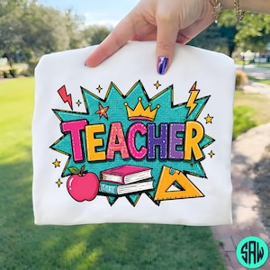 May include: White t-shirt with a vibrant graphic. The design spells out "TEACHER" in pink, yellow, and purple, with a crown, apple, books, and a set square. The shirt is held up by a hand.