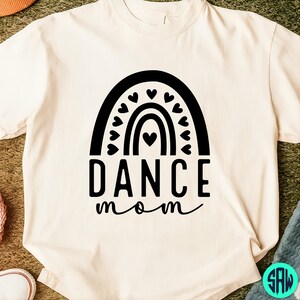May include: Cream-colored t-shirt featuring a black graphic of a rainbow with hearts and the words "DANCE mom" in a bold, stylized font. The design is centered on the front of the shirt, creating a simple yet eye-catching look.