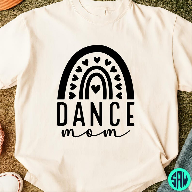 May include: Cream-colored t-shirt featuring a black graphic of a rainbow with hearts and the words "DANCE mom" in a bold, stylized font. The design is centered on the front of the shirt, creating a simple yet eye-catching look.