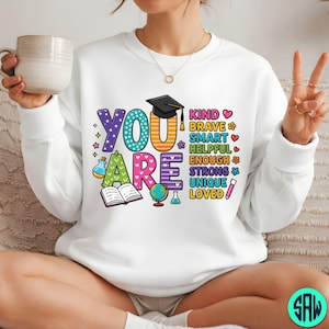 May include: White sweatshirt featuring the words "YOU ARE" in bright, playful lettering. The design includes a graduation cap, an open book, a globe, and other educational symbols, along with positive adjectives.