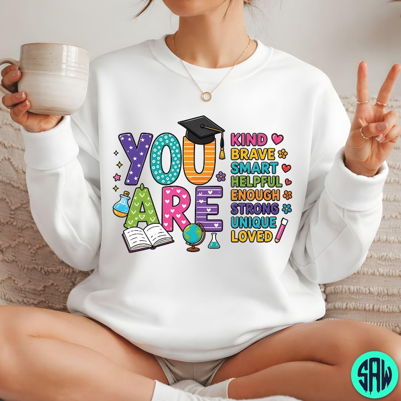 May include: White sweatshirt featuring the words "YOU ARE" in bright, playful lettering. The design includes a graduation cap, an open book, a globe, and other educational symbols, along with positive adjectives.