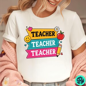 May include: White t-shirt featuring the word "TEACHER" in bold, colourful text. The design includes a yellow banner with a smiley face, a teal banner, and a pink banner with a pencil and a red apple and daisy.