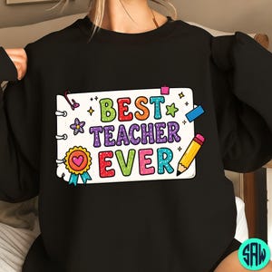 May include: Black sweatshirt featuring the text "BEST TEACHER EVER" in a colourful design. The graphic includes a notepad, pencil, stars, and a heart-shaped award ribbon. The text is in various colours, including green, orange, purple, and red.