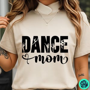May include: A beige t-shirt with the words "DANCE mom" in black, with silhouettes of dancers incorporated into the word "DANCE". The word "mom" is written in a cursive font with a heart symbol. The shirt has a mock neck.