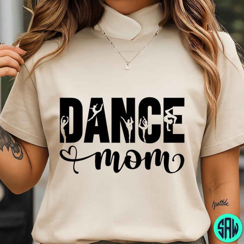 May include: A beige t-shirt with the words "DANCE mom" in black, with silhouettes of dancers incorporated into the word "DANCE". The word "mom" is written in a cursive font with a heart symbol. The shirt has a mock neck.