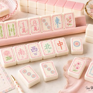 A set of cream-coloured mahjong tiles featuring pink borders and various floral, bamboo, and character designs in green, blue, and red. The tiles are arranged on a light pink wooden rack, with additional loose tiles and a score sheet nearby.