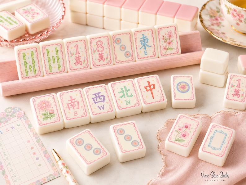 A set of cream-coloured mahjong tiles featuring pink borders and various floral, bamboo, and character designs in green, blue, and red. The tiles are arranged on a light pink wooden rack, with additional loose tiles and a score sheet nearby.