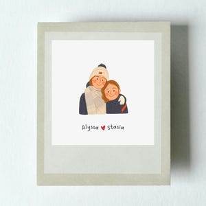 May include: A framed illustration of two people embracing, wearing winter hats and scarves. The text "Alyssa ❤️ stasia" is at the bottom of the image. The artwork is in a polaroid-style frame.