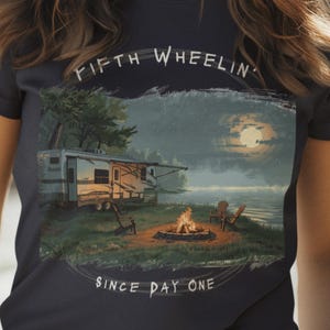 Puede incluir: Camiseta azul marino con un gráfico escénico de una caravana junto a un lago bajo la luna llena, con el texto "FIFTH WHEELIN' SINCE DAY ONE". La camiseta es de color azul oscuro.