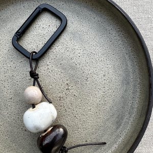 Peut inclure: Un porte-clés composé d'un mousqueton rectangulaire noir mat attaché à un cordon en cuir marron foncé. Le cordon est enfilé à travers trois perles lisses et arrondies de tailles et de couleurs variées : une petite perle beige, une perle blanche plus grande et une perle marron foncé.